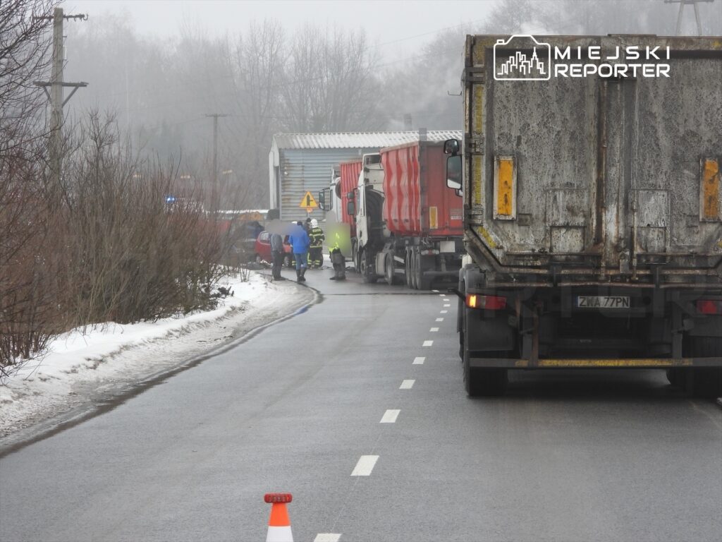 Grupa osób w odzieży roboczej prowadzi działania na drodze, obok zderzonych ciężarówek w mglistym otoczeniu.