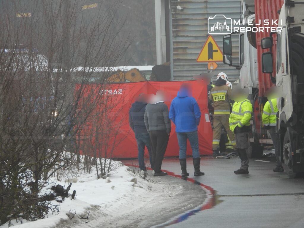 Grupa osób w odzieży roboczej oraz cywilnej zbiera się wokół miejsca zdarzenia drogowego, gdzie pracują strażacy.