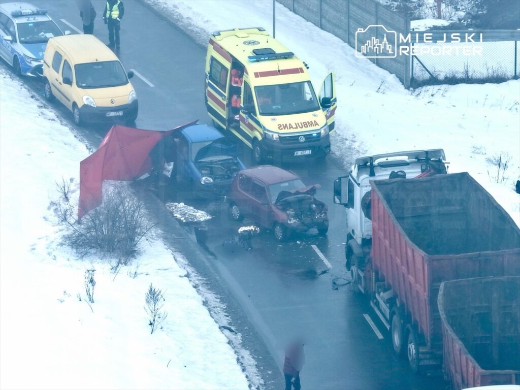Na drodze znajduje się wypadek samochodowy; niebieski i czerwony samochód z uszkodzeniami, obok karetka i policyjny radiowóz.