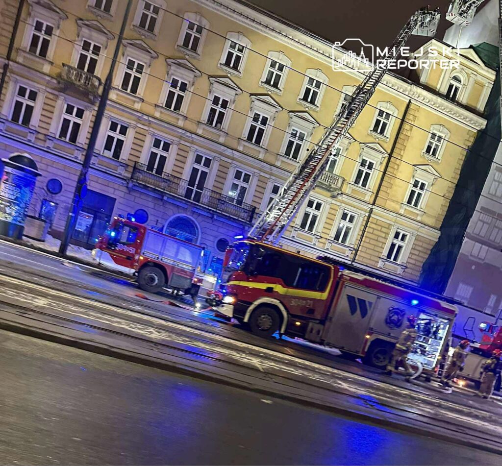 Wozy strażackie z włączonymi sygnałami świetlnymi stoją na ulicy przed kamienicą w nocy.