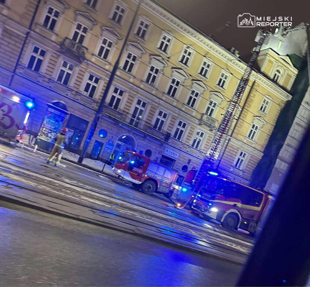 Strażacy w mundurach zabezpieczają teren akcji ratunkowej przy oświetlonym budynku w nocy.