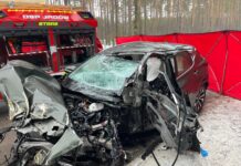 Zniszczony samochód osobowy leży na drodze, otoczony przez czerwone namioty służb ratunkowych i drzewami w tle.