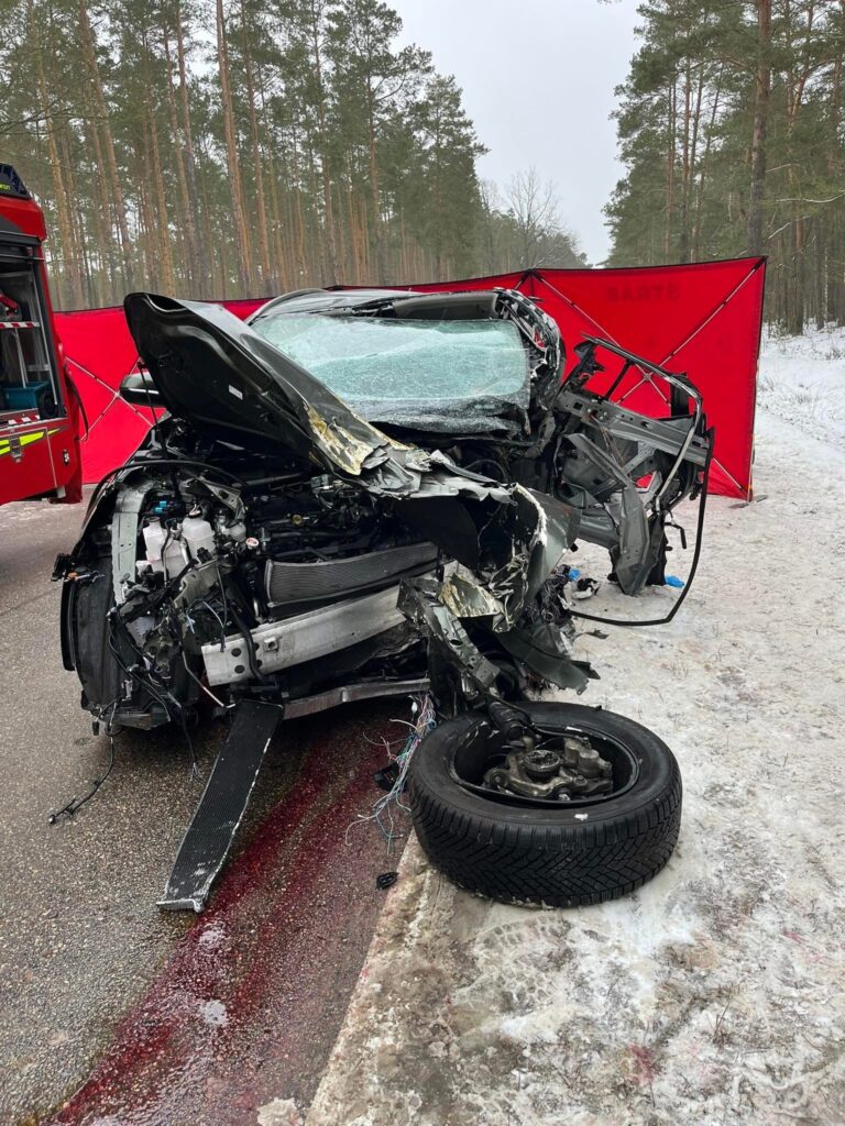 Zniszczony samochód osobowy leży na poboczu drogi, otoczony przez sosnowy las, z widocznymi uszkodzeniami przodu.