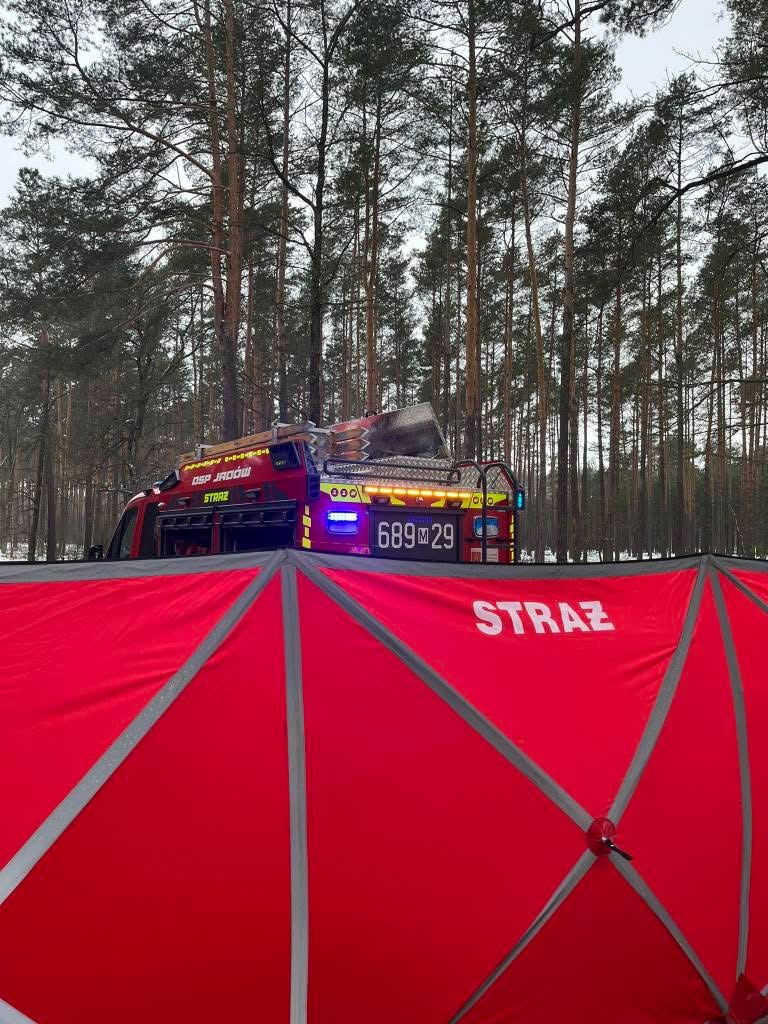 W tle gęstego lasu, widać wóz strażacki z włączonymi sygnałami świetlnymi, a przed nim rozłożony czerwony namiot z napisem "STRAŻ".