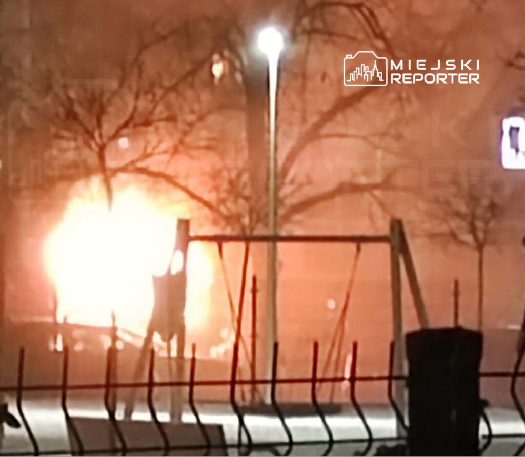 Płonący pojazd na osiedlu, w tle widać oświetlone drzewa i plac zabaw.