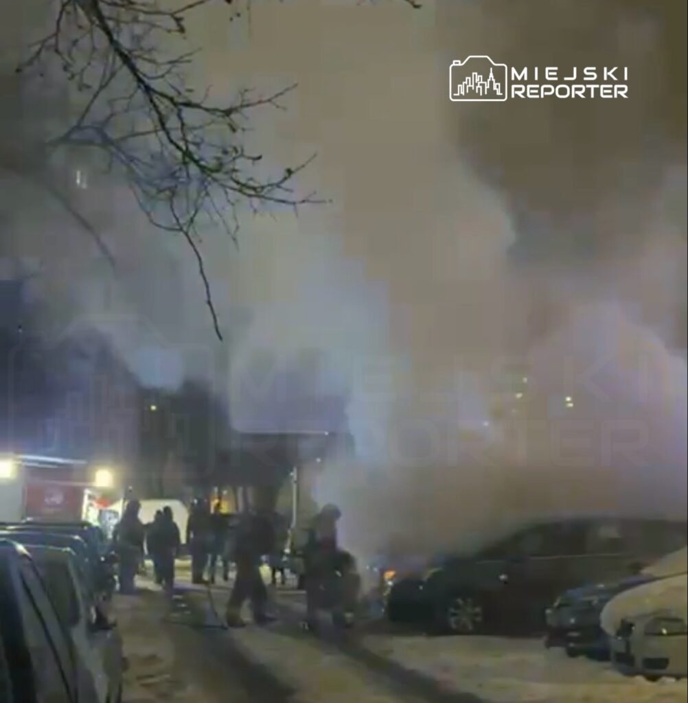 Grupa strażaków w mundurach gasząca pożar w pobliżu zaparkowanych samochodów w oświetlonym śniegiem otoczeniu.