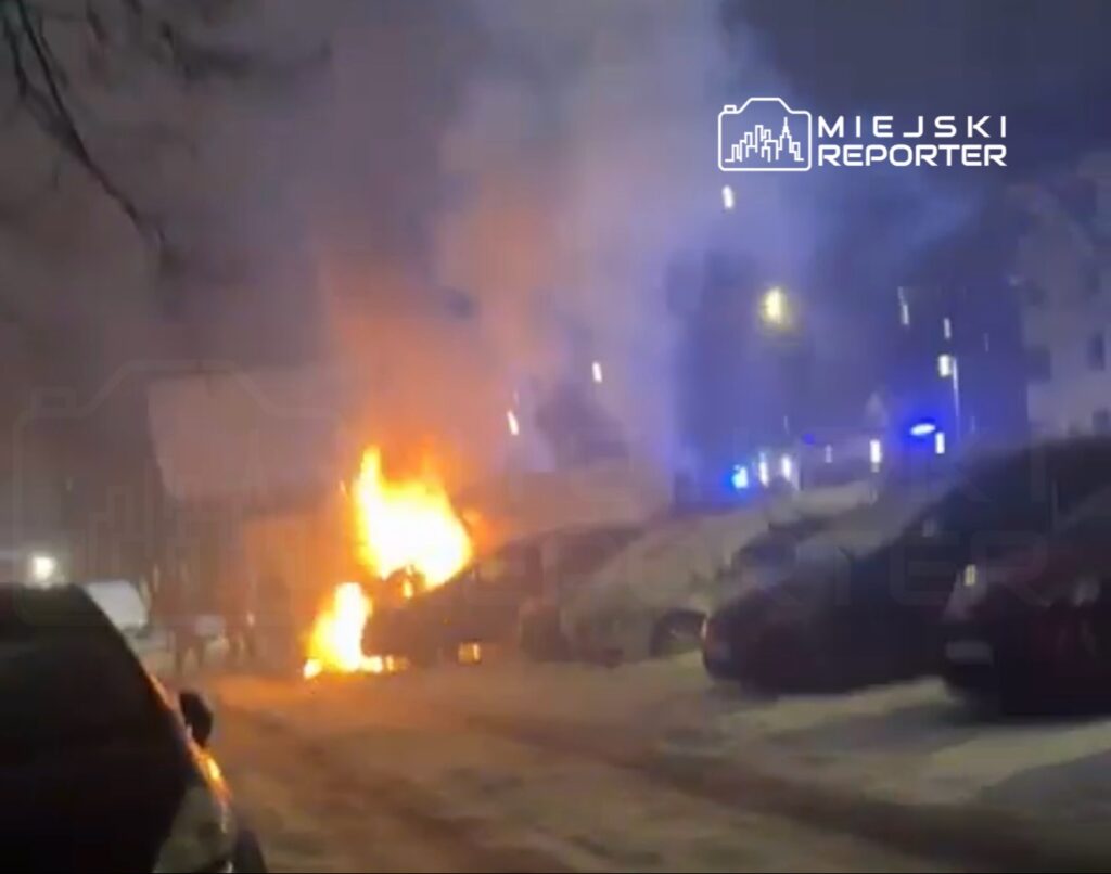 Ogień wydobywający się z zaparkowanego samochodu w zaśnieżonym otoczeniu, w tle widoczne niebieskie światła służb.