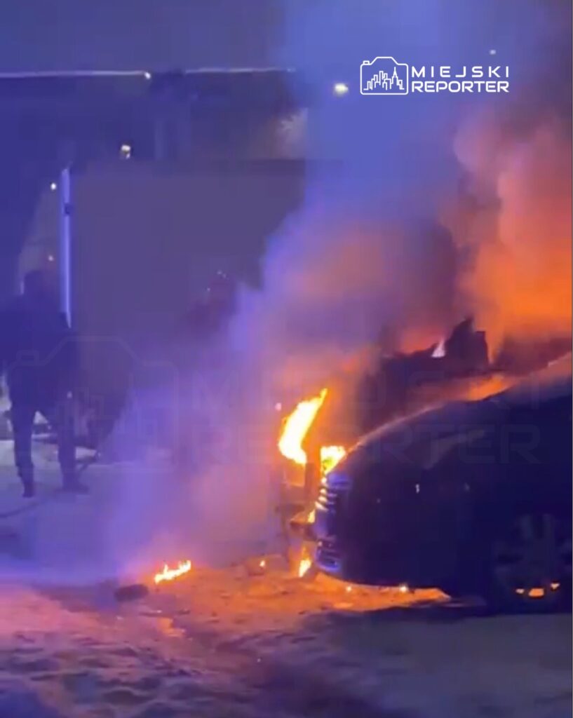 Płonący samochód na parkingu, otoczony dymem i ogniem, w nocy.