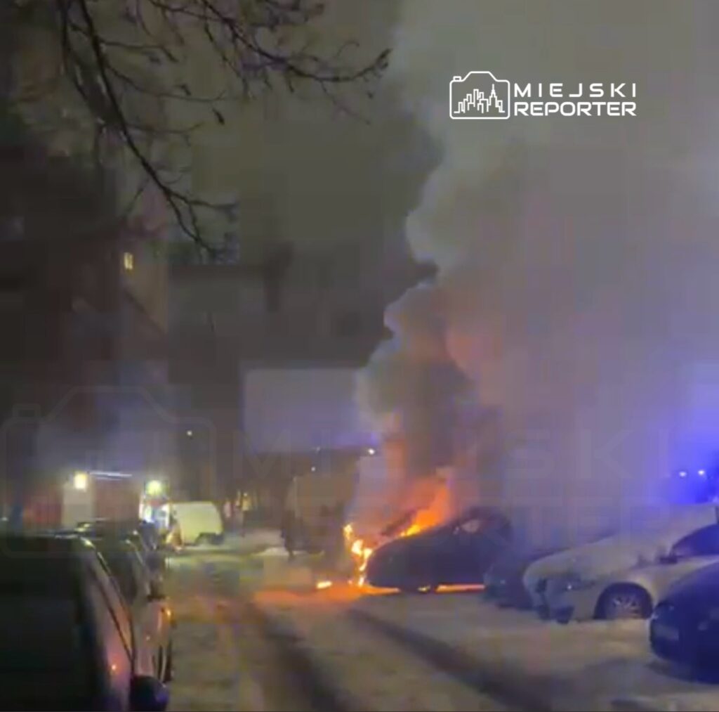 Samochód płonący na parkingu, otoczony przez gęsty dym, w tle widać policyjne światła i inne zaparkowane pojazdy.