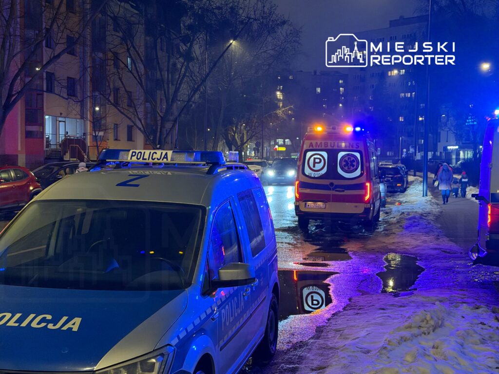 Policyjny radiowóz z włączonymi sygnałami świetlnymi stoi na ulicy, obok karetki pogotowia.