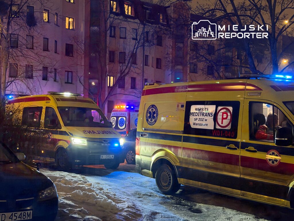 Dwa karetki pogotowia z włączonymi sygnałami świetlnymi zaparkowane na ośnieżonej ulicy w pobliżu budynku mieszkalnego.