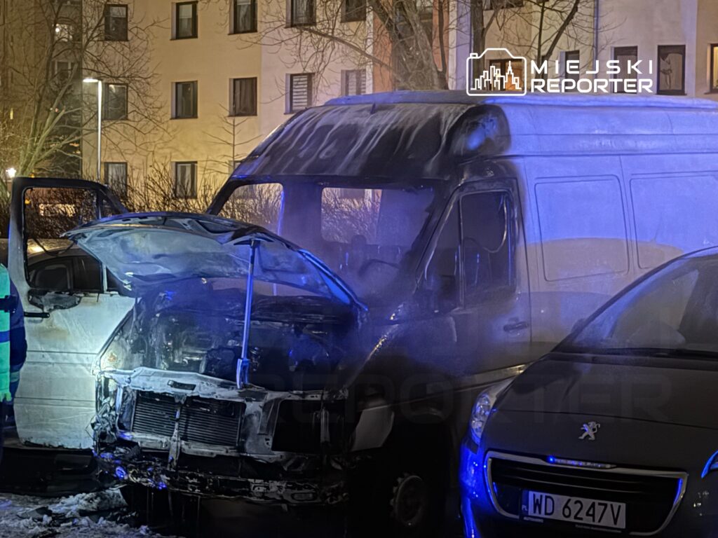 Samochód dostawczy z uszkodzoną przednią częścią, obok stoi strażak w mundurze, a w tle widać bloki mieszkalne.