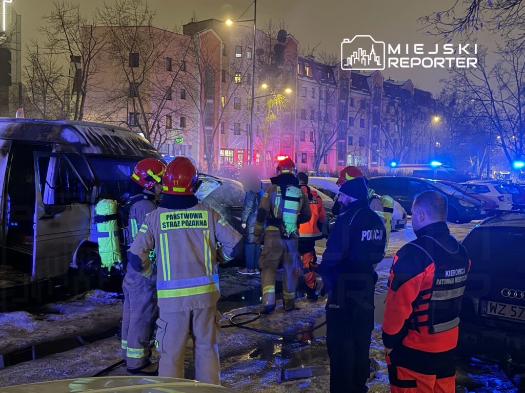 Grupa strażaków w hełmach i mundurach pracuje przy uszkodzonym pojeździe, otoczona przez policyjnych funkcjonariuszy i ratowników medycznych.