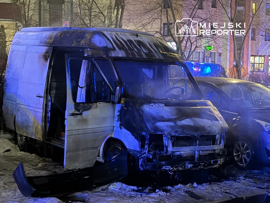 Spalony furgon z otwartymi drzwiami, obok stoi uszkodzony samochód, w tle migające niebieskie światła.