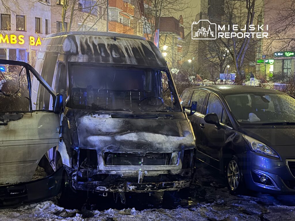 Spalony van z otwartymi drzwiami stoi na śniegu obok nieuszkodzonego samochodu osobowego w miejskim otoczeniu.