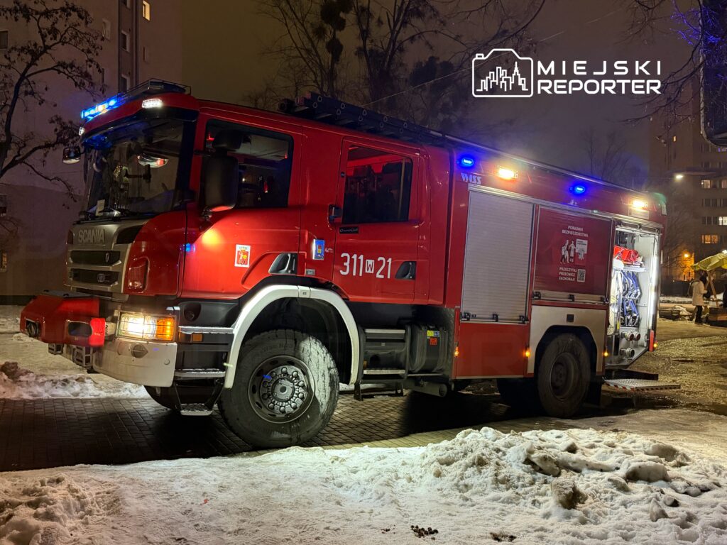 Czerwony wóz strażacki z włączonymi sygnałami świetlnymi stoi na ośnieżonym placu w nocy.