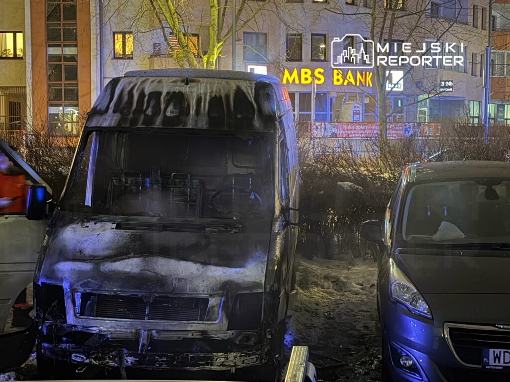 Spalony furgon zaparkowany na chodniku, obok niego stoi drugi samochód osobowy, w tle widoczny neon banku.