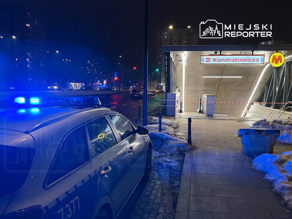 Policyjny radiowóz z włączonymi sygnałami świetlnymi stoi przy stacji metra Kondratowicza, w tle widoczne oświetlenie uliczne.