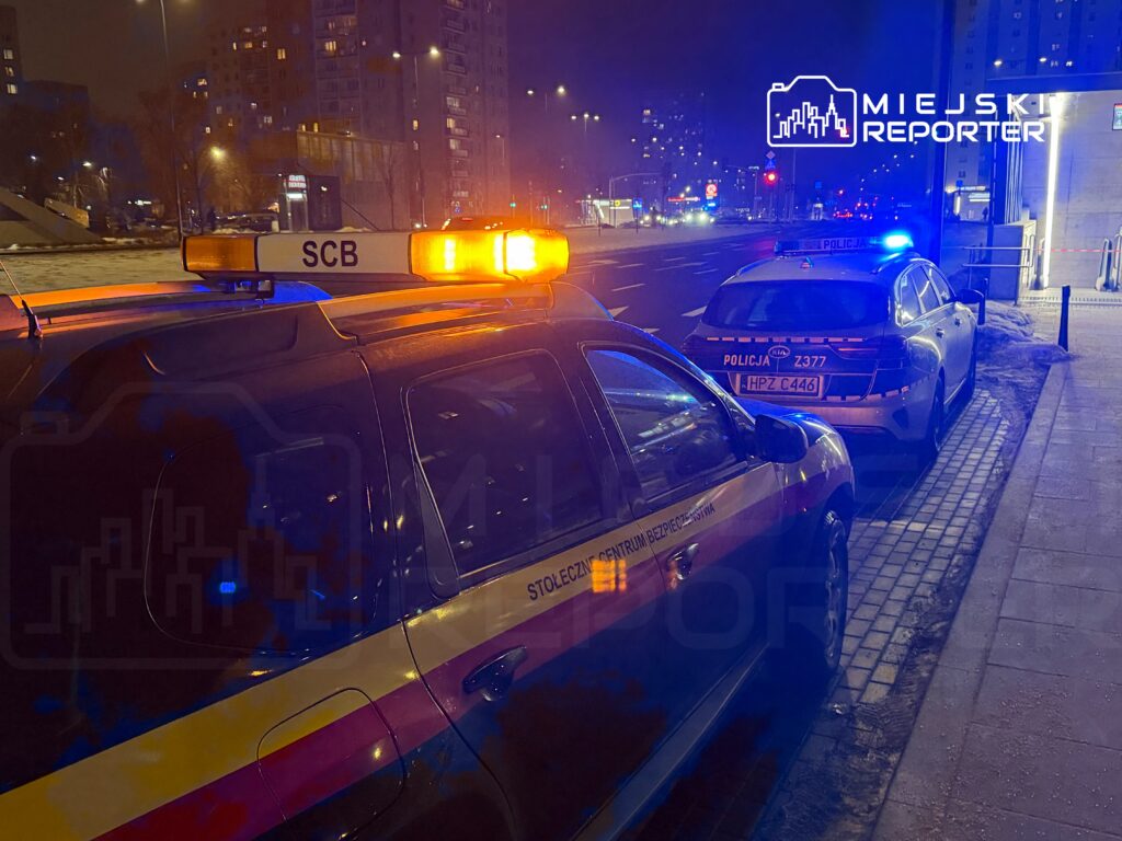 Radiowóz policyjny z włączonymi sygnałami świetlnymi stoi na ulicy w nocy, obok innego pojazdu służbowego.