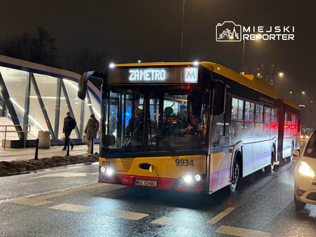 Autobus z wyświetlaczem "ZAMETRO" zatrzymuje się na przystanku, obok przechodzą piesi w ciemnej, nocnej scenerii.