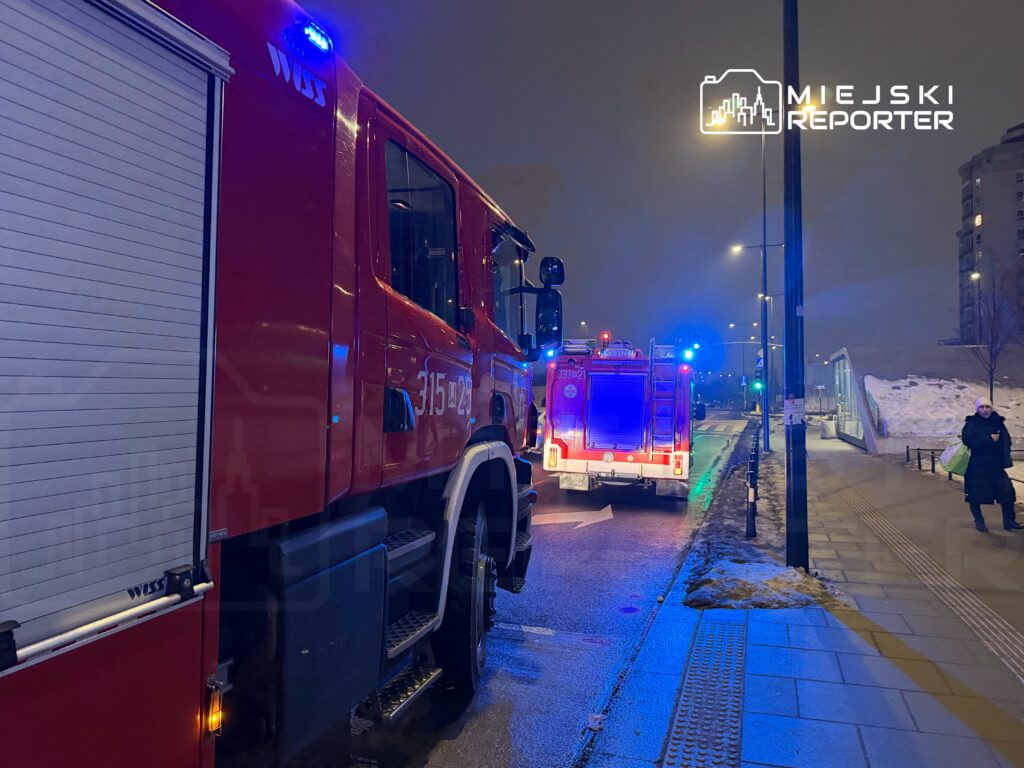 Czerwony wóz strażacki z włączonymi sygnałami świetlnymi stoi na ulicy, obok drugiego wozu strażackiego.