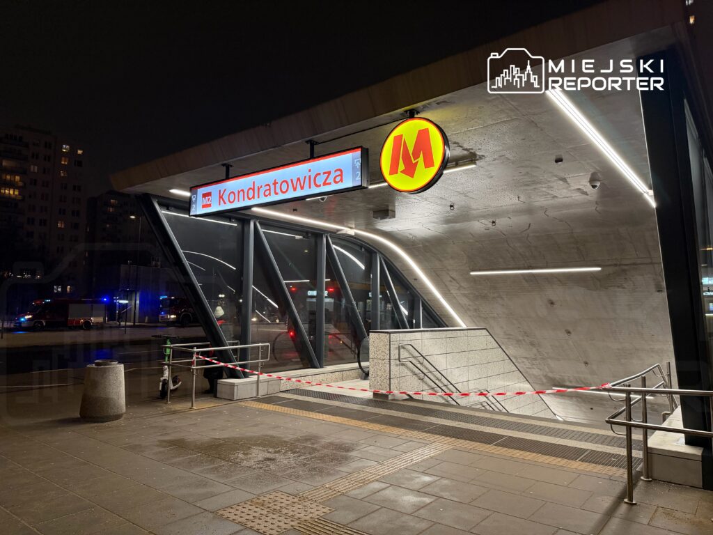 Stacja metra Kondratowicza z zamkniętym wejściem, otoczona taśmą ostrzegawczą, w nocy z widocznymi pojazdami służb ratunkowych.
