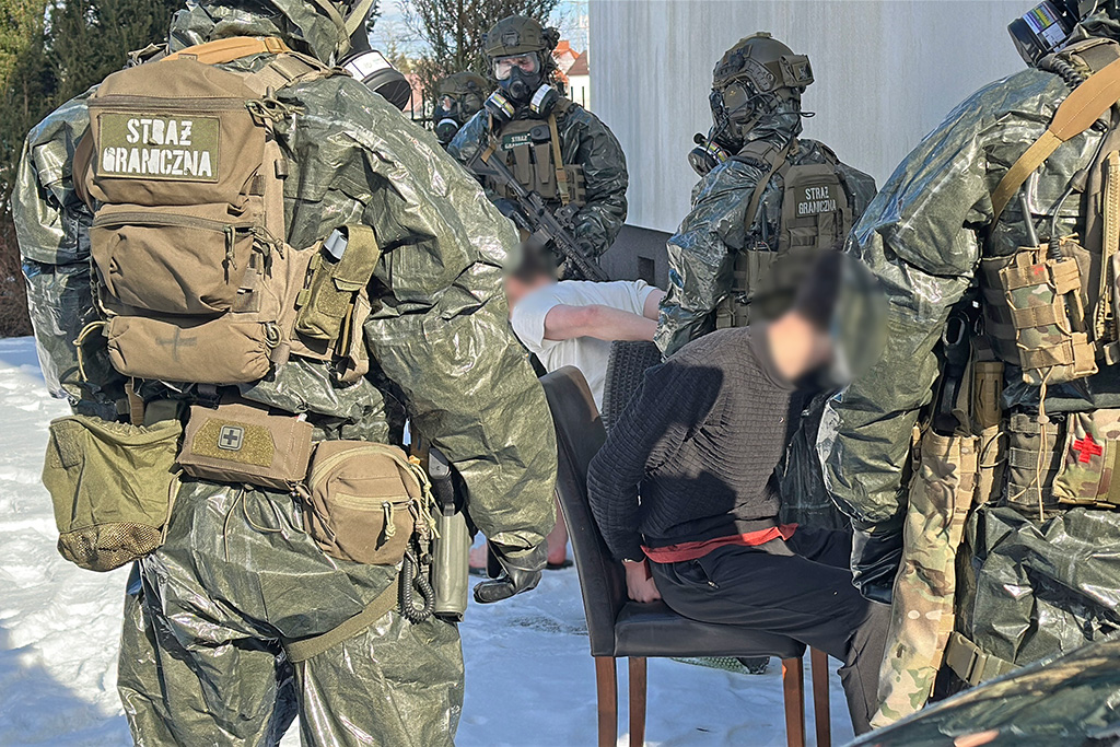 Grupa funkcjonariuszy w mundurach, wyposażonych w sprzęt ochronny, otacza mężczyznę siedzącego na krześle.