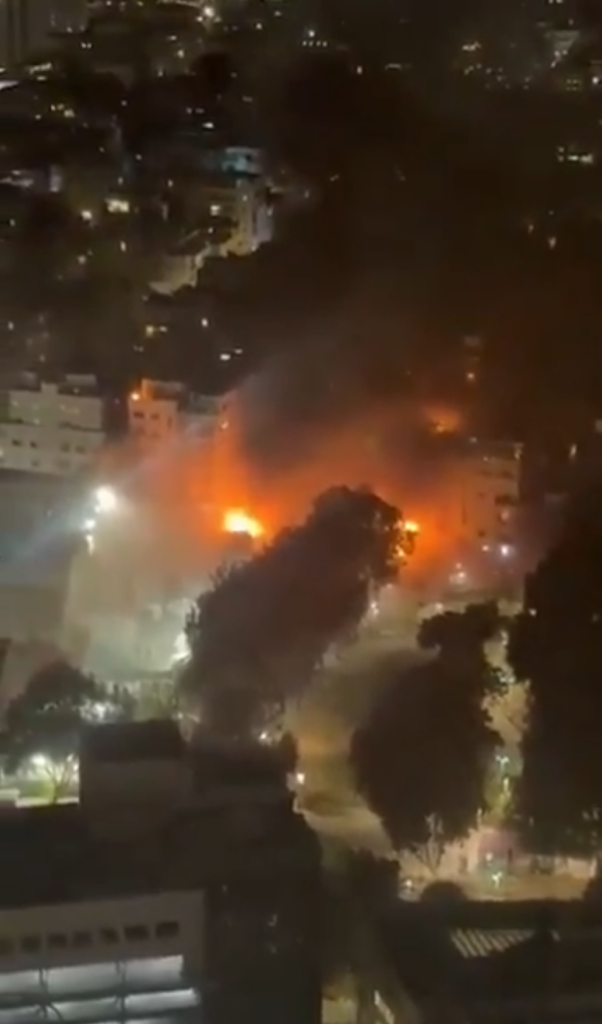 W nocy widać ogień i dym unoszący się z budynku w miejskim otoczeniu, oświetlonym przez latarnie.