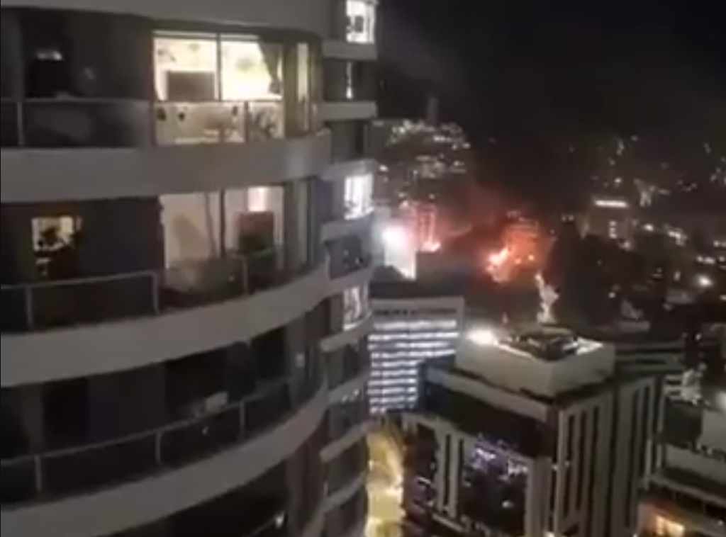 Widok z wysokiego budynku na nocne miasto, z oświetlonymi oknami oraz migającymi światłami w tle.