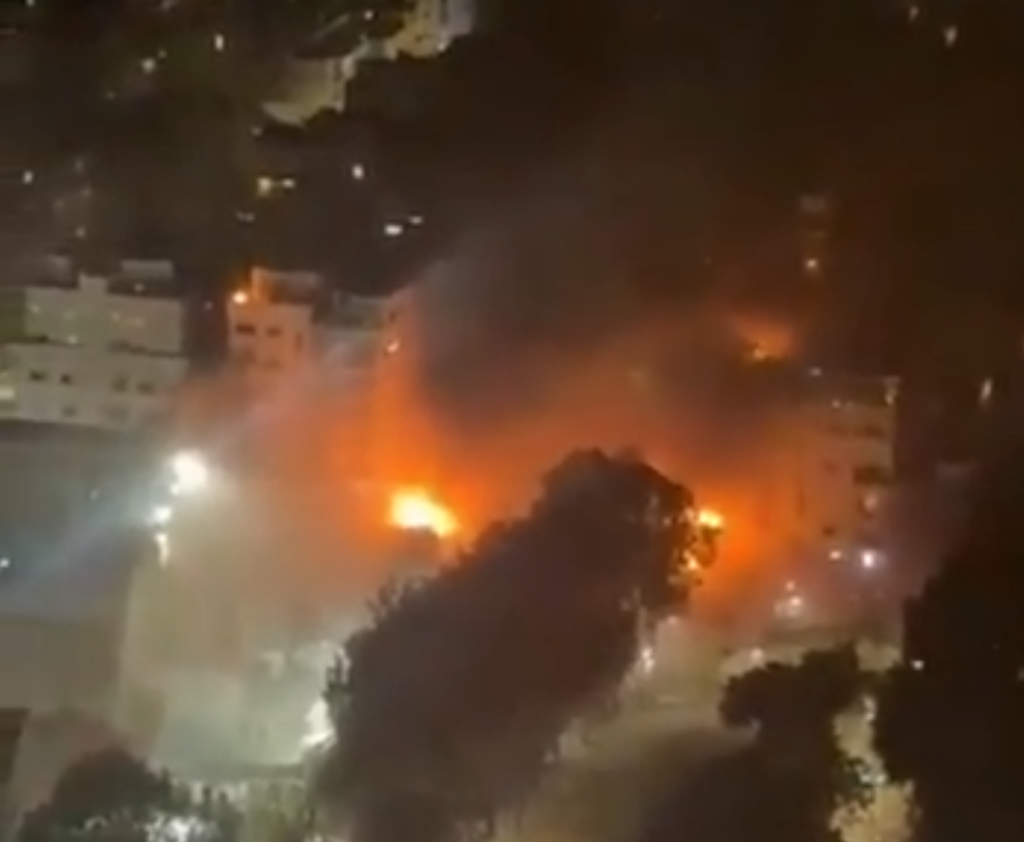 Wysoka chmura dymu i ogień wydobywający się z budynku w nocy, otoczonego przez inne oświetlone budynki.