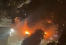 Wysoka chmura dymu i ogień wydobywający się z budynku w nocy, otoczonego przez inne oświetlone budynki.