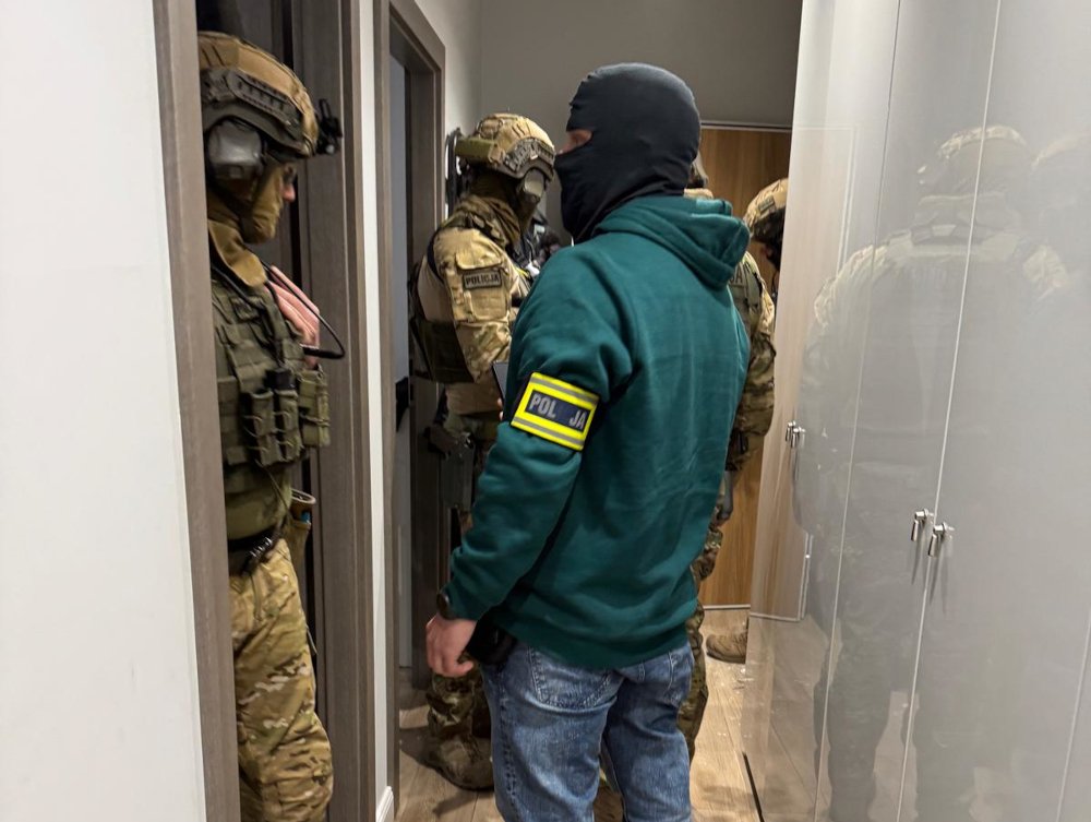 Mężczyzna w zielonym dresie z oznakowaniem "POLICJA" stoi obok uzbrojonych funkcjonariuszy w mundurach, w pomieszczeniu.