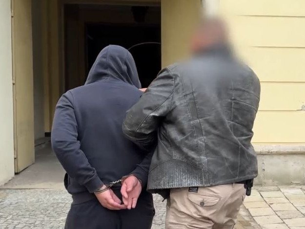 Mężczyzna w dresie z założoną kapturem jest prowadzony przez policjanta w skórzanej kurtce.