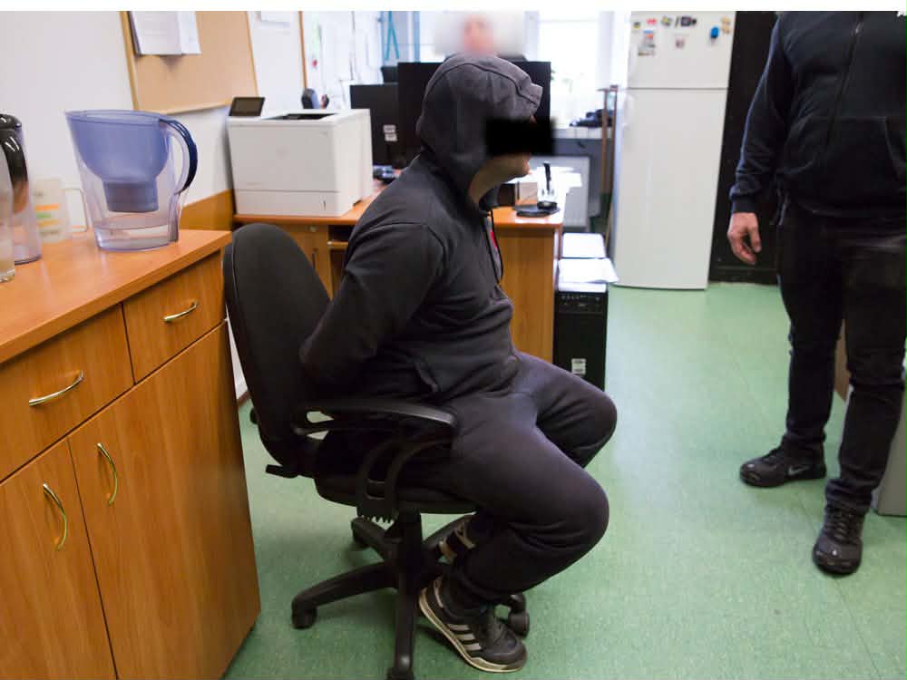 Mężczyzna w czarnej bluzie z kapturem siedzi na krześle obrotowym z rękami związanymi, w biurze z drewnianymi meblami.