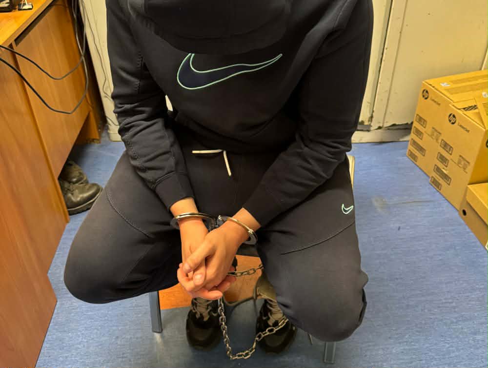 Młody mężczyzna w dresie siedzi na krześle z rękami skuty w kajdankach, w pomieszczeniu z biurowym wyposażeniem.