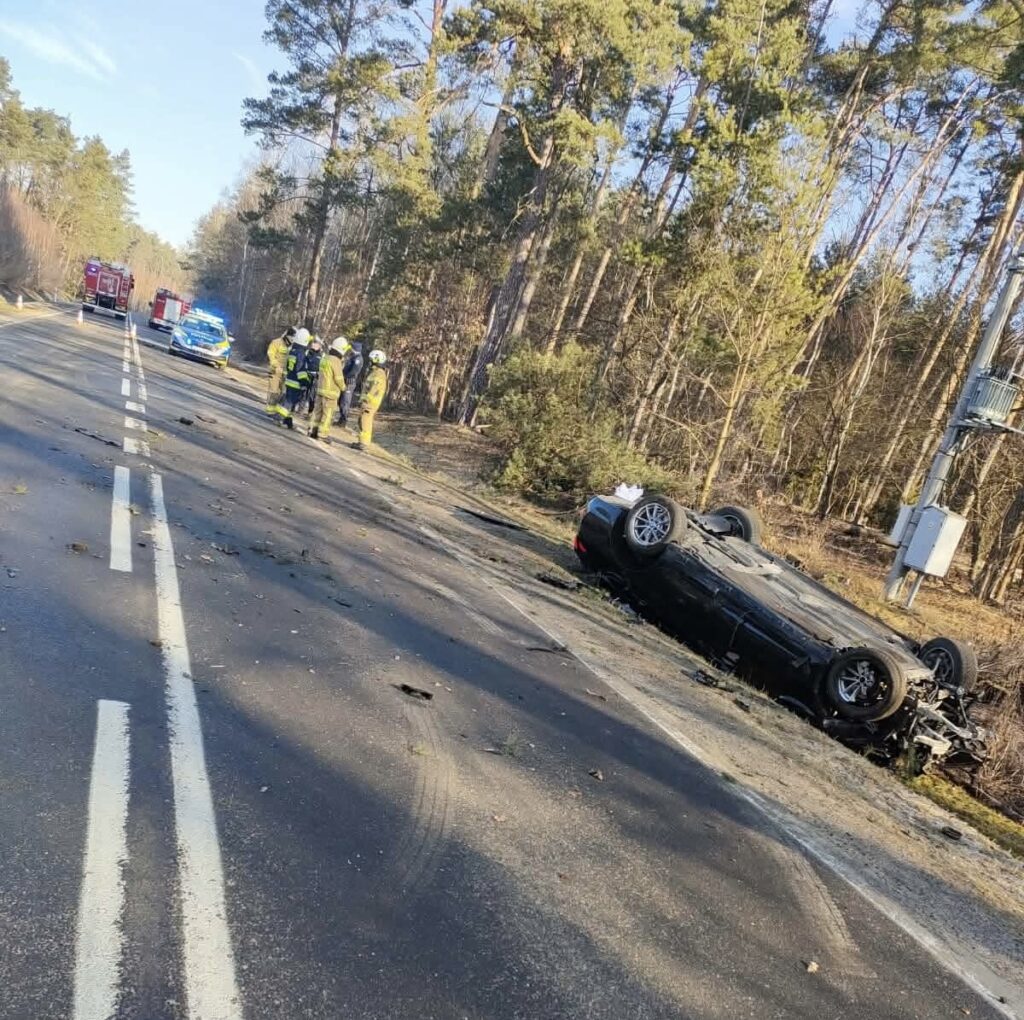 Na drodze leży przewrócony samochód osobowy, a obok grupka strażaków i policjantów prowadzi akcję ratunkową. W tle widać las.