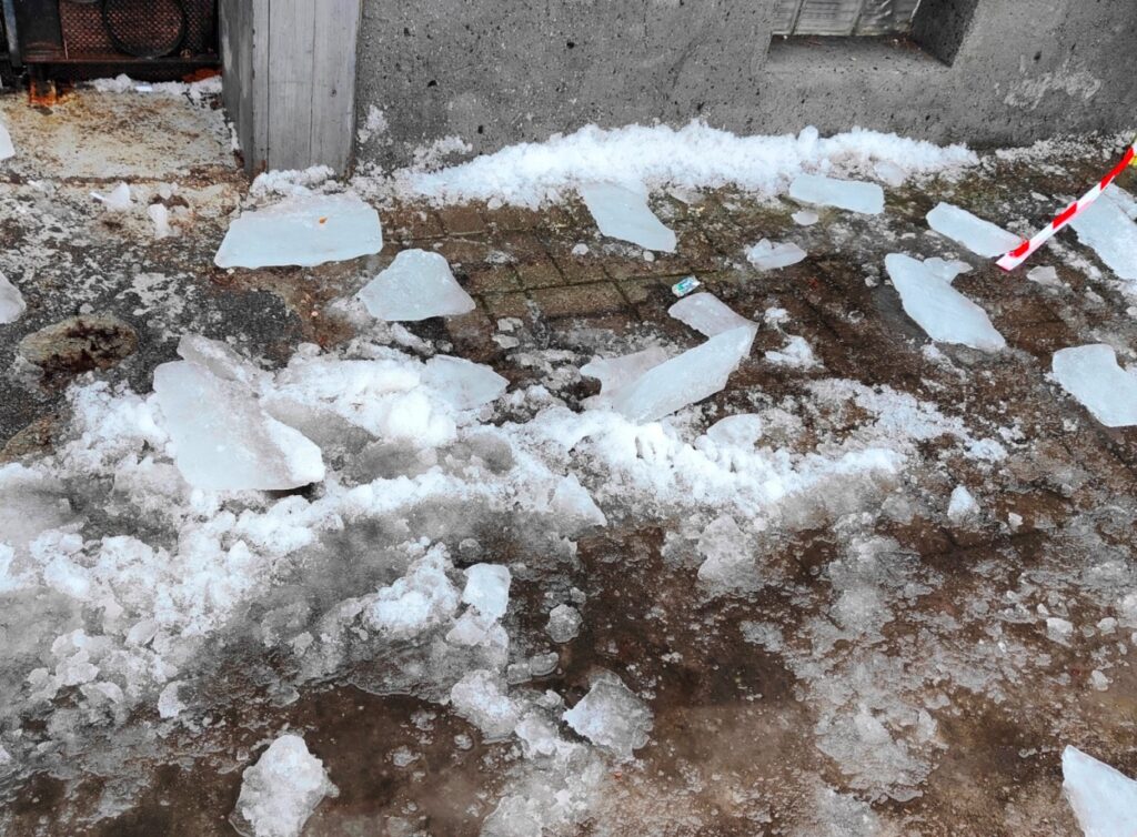 Rozbite kawałki lodu i śniegu leżą na chodniku obok szarej ściany budynku, tworząc niebezpieczne śliskie miejsce.