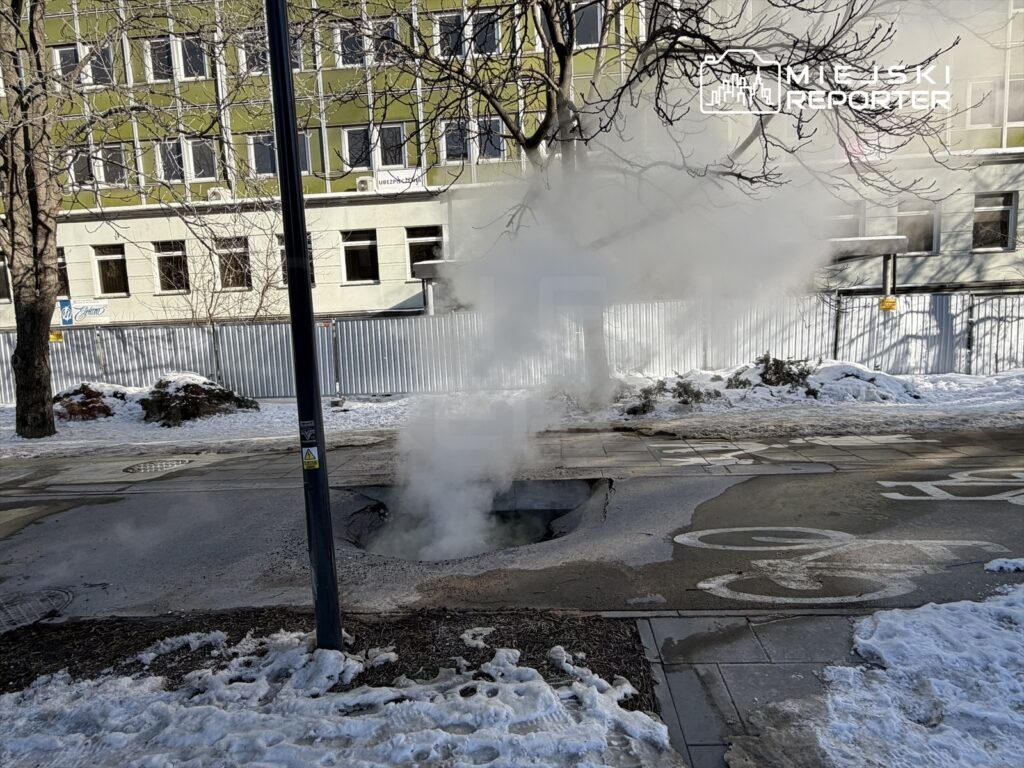 Para wydobywająca się z otworu w jezdni w zimowej scenerii, w tle widoczny budynek z przeszkloną fasadą.