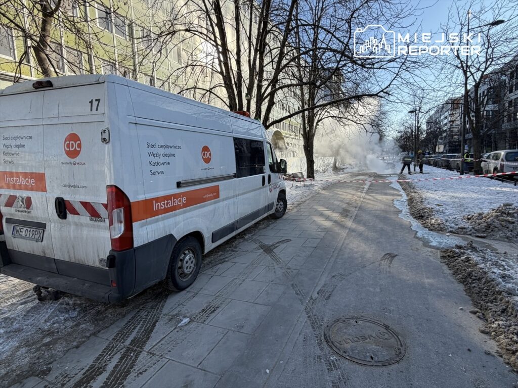 Pracownicy w kamizelkach odblaskowych usuwają awarię rury ciepłowniczej obok zaparkowanego pojazdu serwisowego.