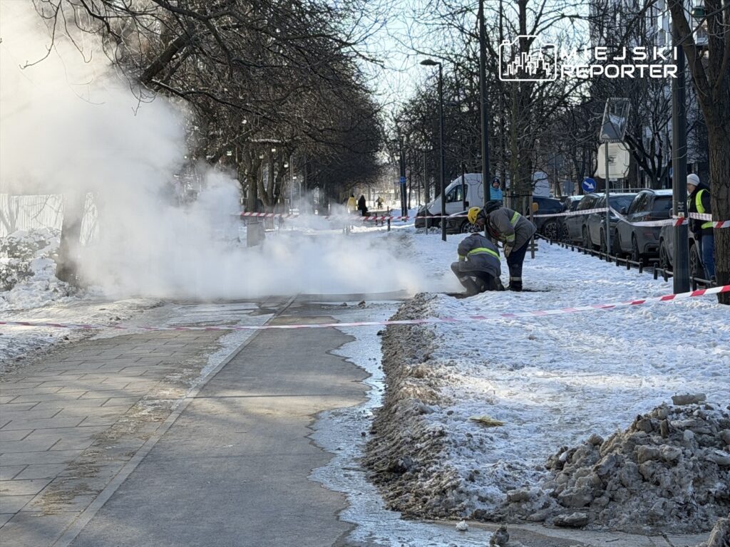 Pracownicy w odblaskowych kamizelkach naprawiają rury w zasypanej śniegiem ulicy, z której wydobywa się para.