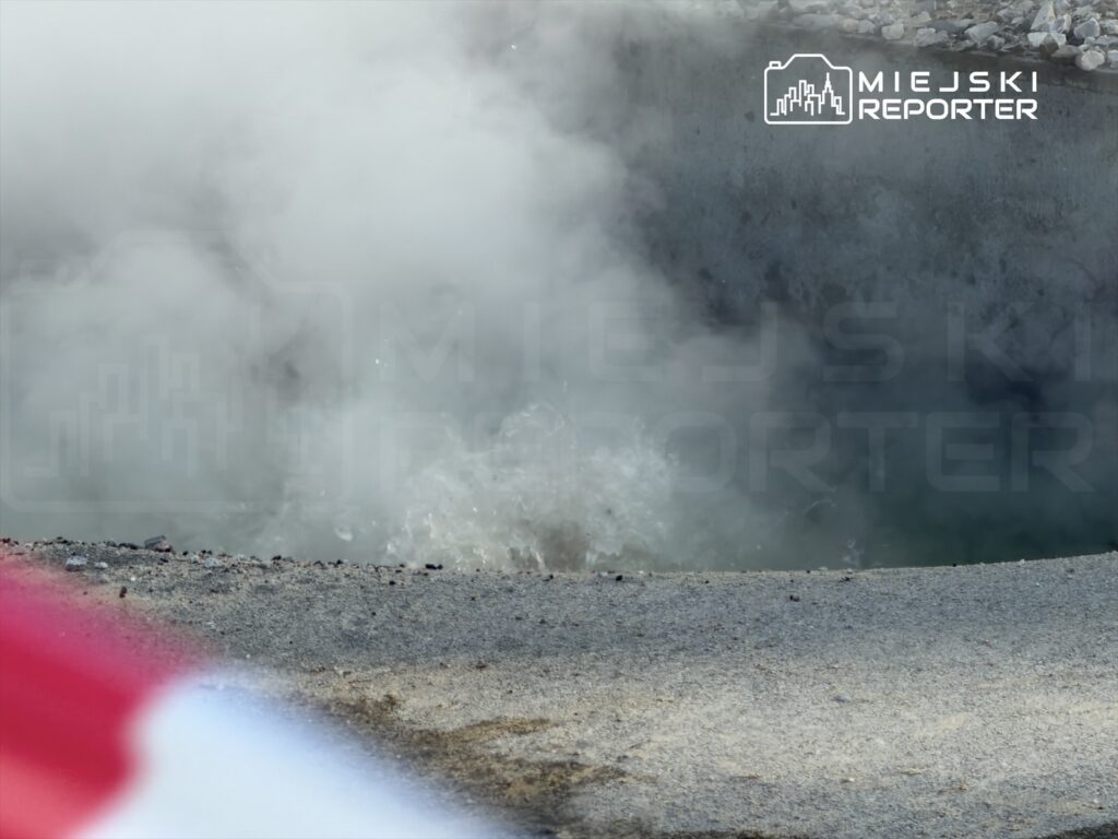 Wysoka para unosząca się nad kraterem, z którego wydobywa się woda, otoczona kamieniami na szarej nawierzchni.