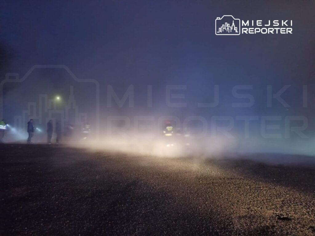 Grupa strażaków pracuje w gęstej mgle na oświetlonej ulicy w nocy.