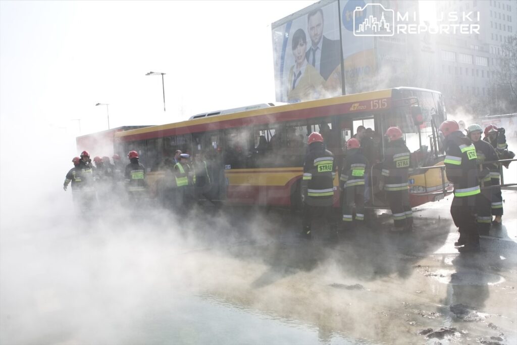 Grupa strażaków w hełmach i mundurach pracuje przy miejskim autobusie, z którego wydobywa się para.