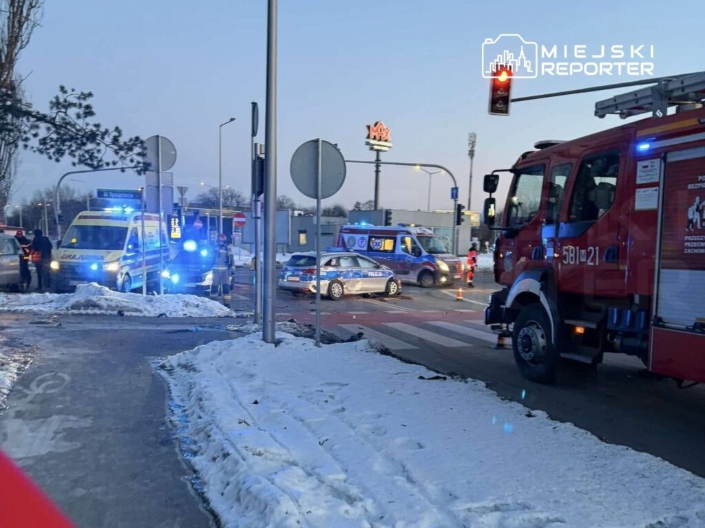 Karetką pogotowia i radiowozami policji zabezpieczona ulica po wypadku, w tle widoczny sklep.