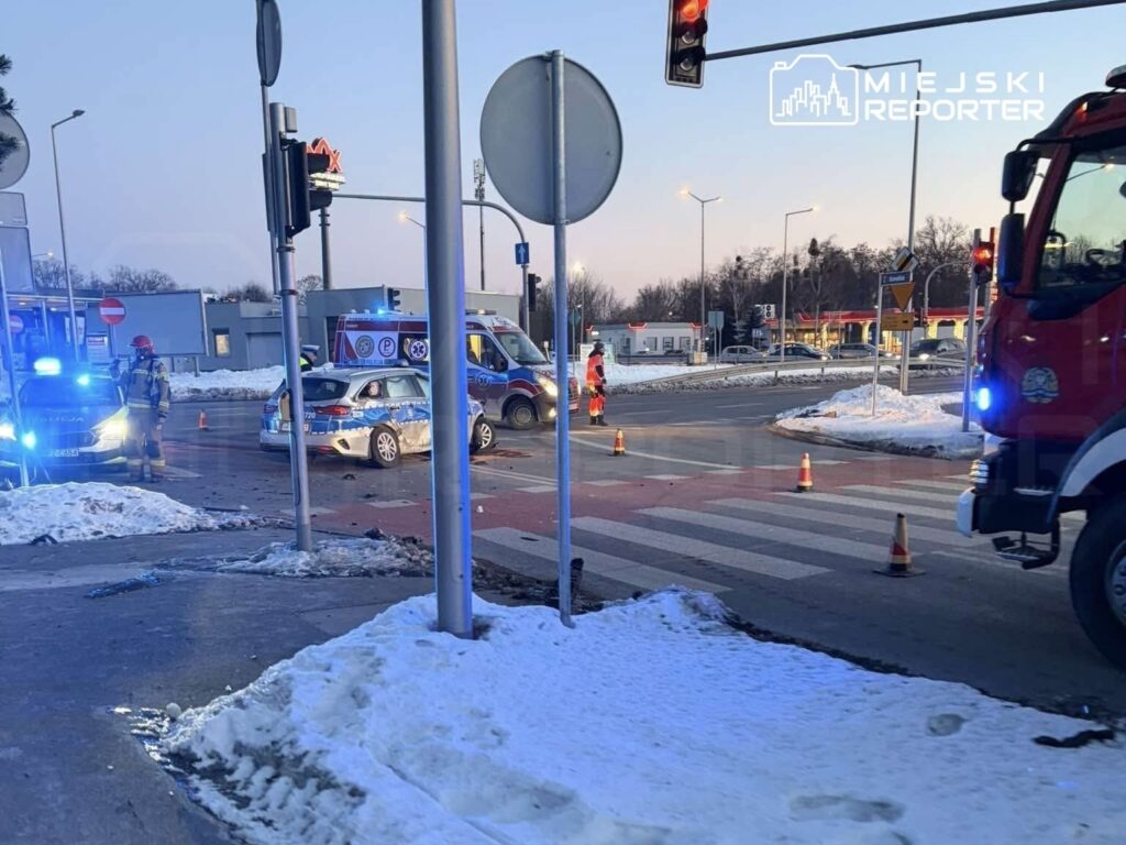 Na skrzyżowaniu z sygnalizacją świetlną interweniują strażacy i policja, wokół uszkodzone pojazdy oraz leżące na jezdni fragmenty.