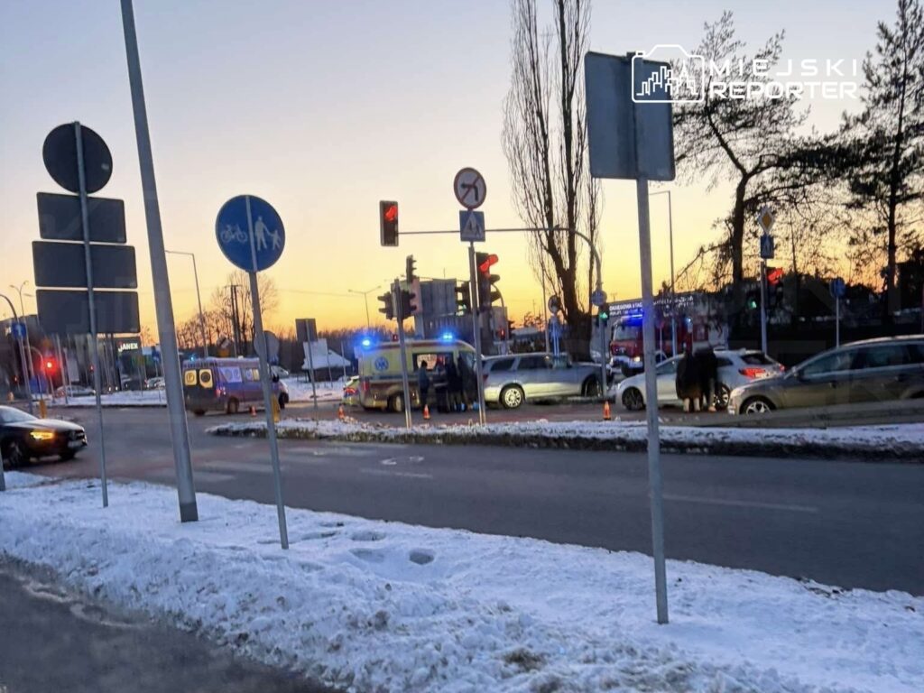 Na skrzyżowaniu z sygnalizacją świetlną stoją pojazdy służb ratunkowych, a w tle widać zachód słońca.