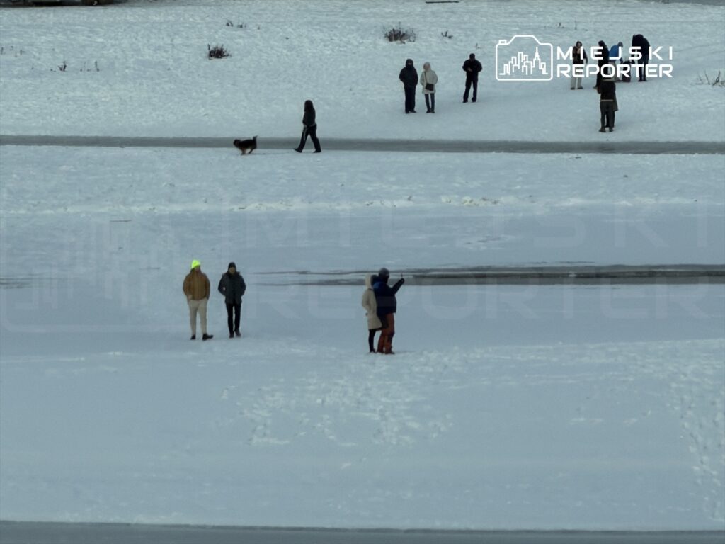 Grupa osób spaceruje po zaśnieżonym terenie, a w tle widoczny jest pies na smyczy.