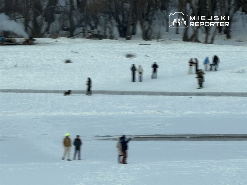 Grupa osób spaceruje po zamarzniętej rzece, a w tle widoczne drzewa i unoszący się dym.