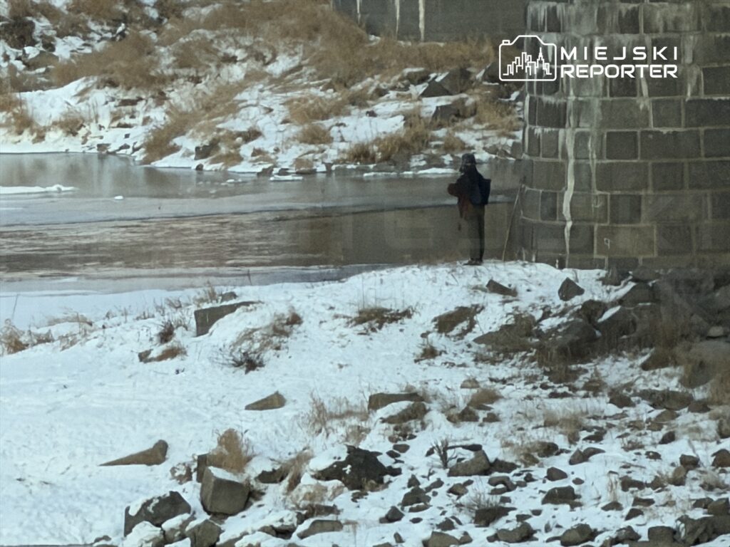 Mężczyzna w zimowej odzieży stojący nad zamarzniętym brzegiem rzeki, obok kamiennego muru.