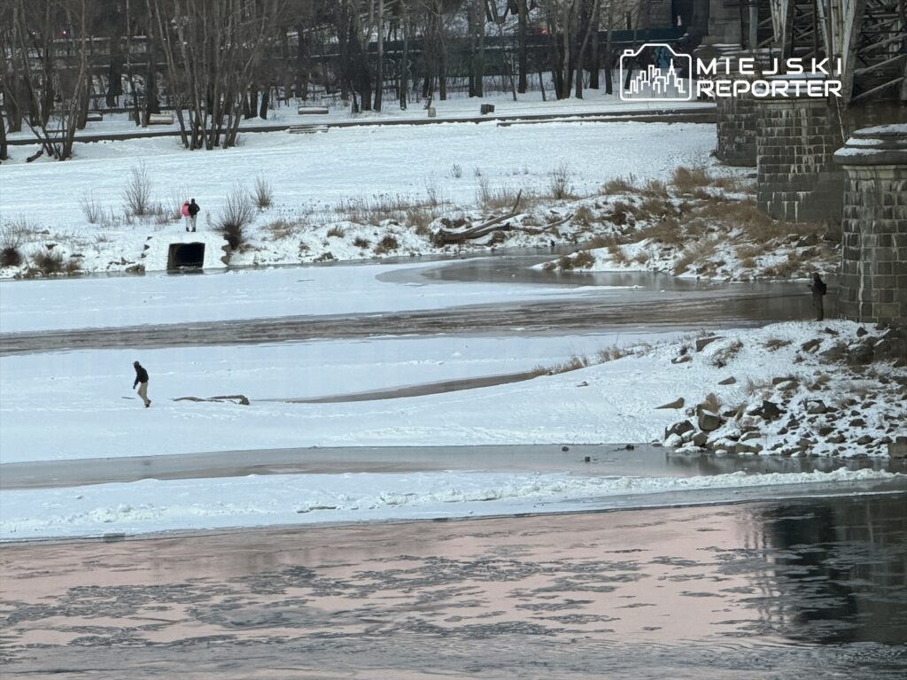 Mężczyzna spacerujący po zamarzniętej rzece, w tle dwie osoby na brzegu w zimowym otoczeniu.